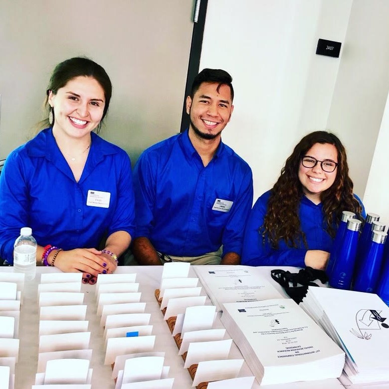 Three students in blue shirts behind a table with nametags for a conference.