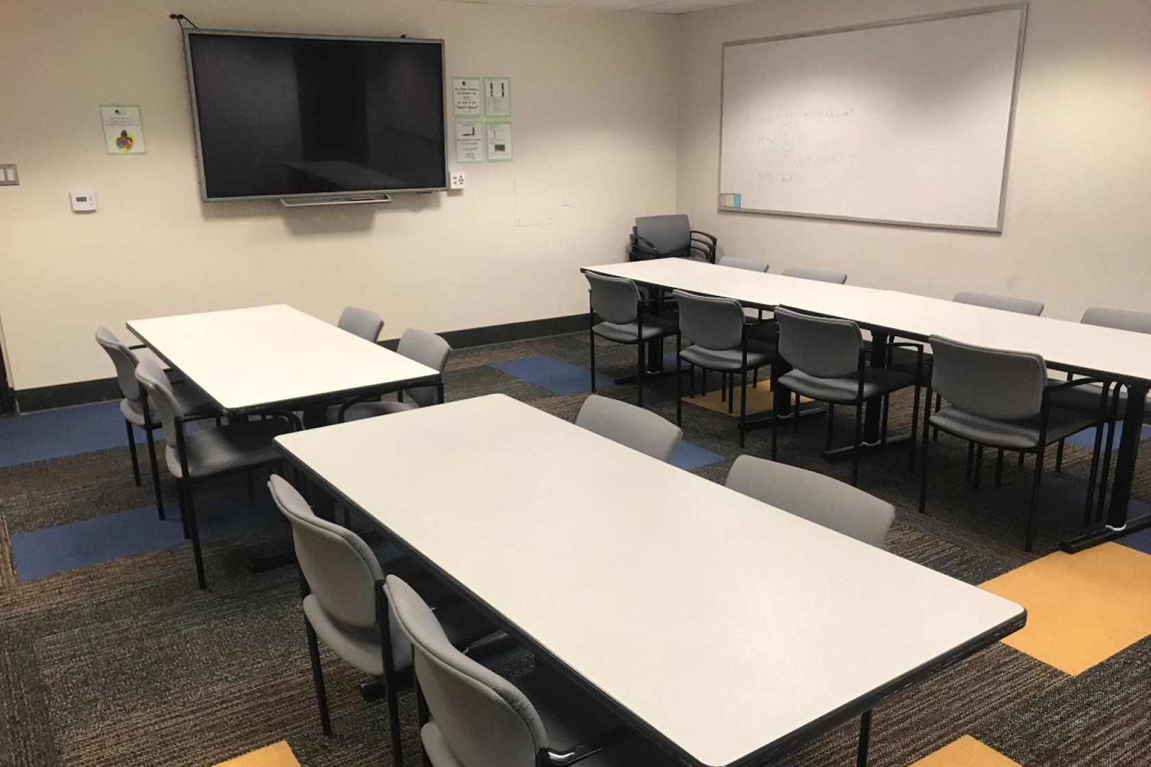 A classroom with a big screen on one wall and a white board on the other wall. There are four long classroom tables with four chairs each.
