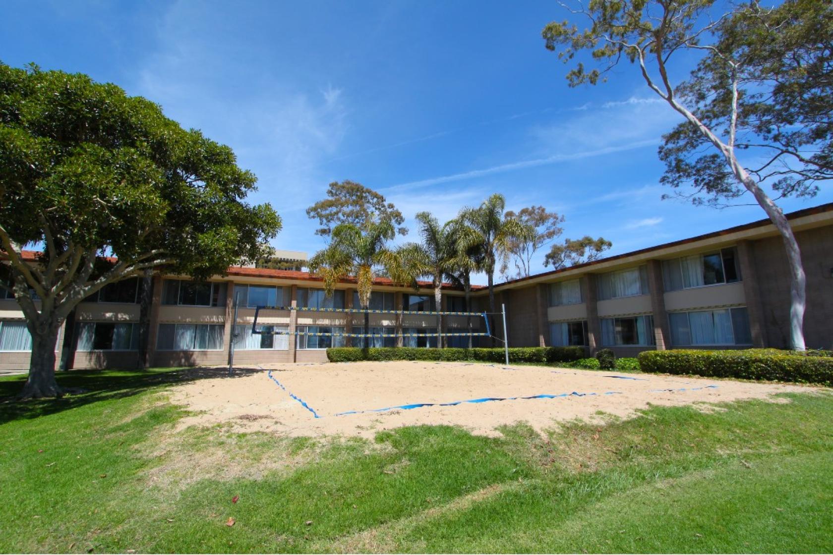 In the foreground there is a green lawn. ON the left there is a big shady tree next to which you can see a sandy beach volleyball court. In the back is a midcentury two-story cinder block residence hall. You can see a sunny and blue sky.